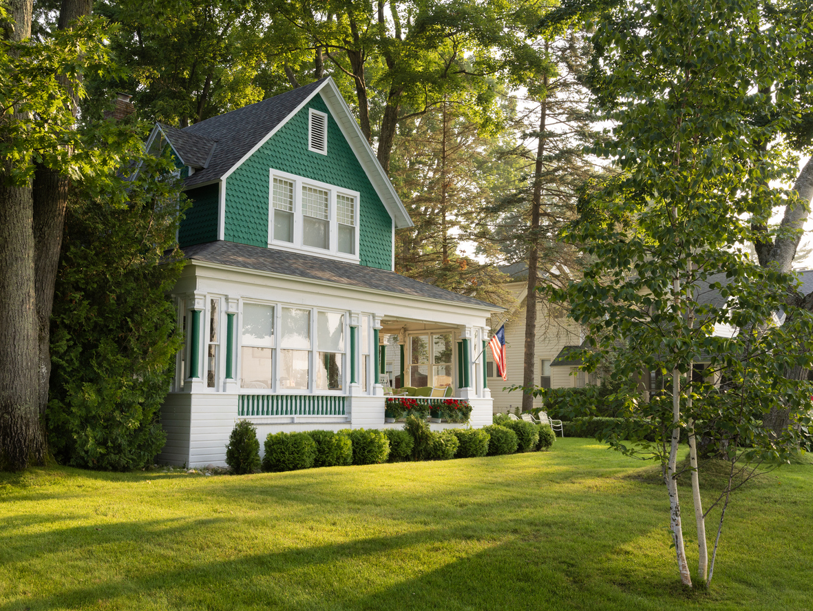 Charming green house with veranda and American flag in sunny woodland setting.